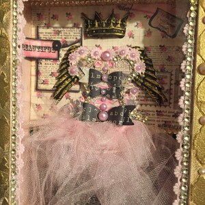 May include: A pink and gold shadow box with a crown, wings, and a tutu. The box has a pink and white floral background with the words "It is a heavy beautiful" and "To things I will never forget".