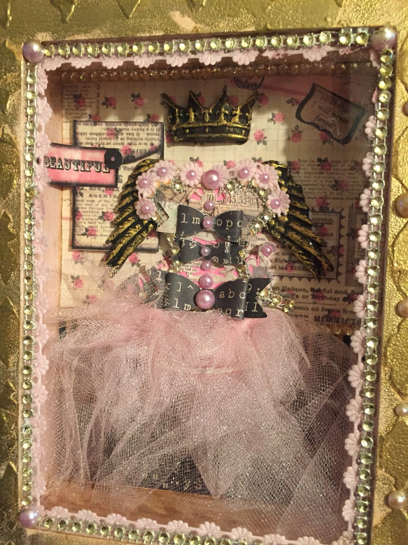 May include: A pink and gold shadow box with a crown, wings, and a tutu. The box has a pink and white floral background with the words "It is a heavy beautiful" and "To things I will never forget".