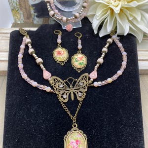 May include: A jewellery set featuring a necklace with a butterfly pendant, a matching bracelet, and earrings. The necklace has a double strand design with pink beads and a butterfly pendant. The earrings and pendant feature floral designs.