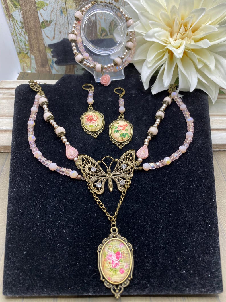May include: A jewellery set featuring a necklace with a butterfly pendant, a matching bracelet, and earrings. The necklace has a double strand design with pink beads and a butterfly pendant. The earrings and pendant feature floral designs.
