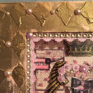May include: A gold frame with a pink and white patterned background. The frame is decorated with rhinestones and pearls. The frame contains a variety of paper crafts, including a crown, a feather, and a paper flower. The text "TIFUL" is visible in the image.