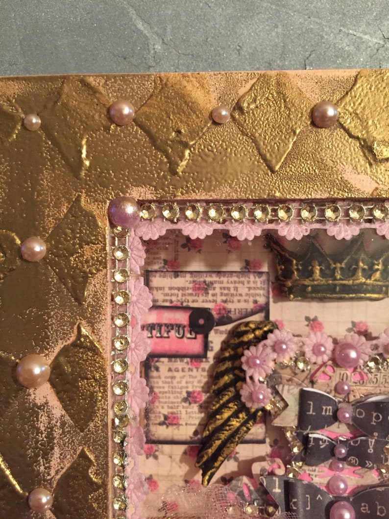 May include: A gold frame with a pink and white patterned background. The frame is decorated with rhinestones and pearls. The frame contains a variety of paper crafts, including a crown, a feather, and a paper flower. The text "TIFUL" is visible in the image.