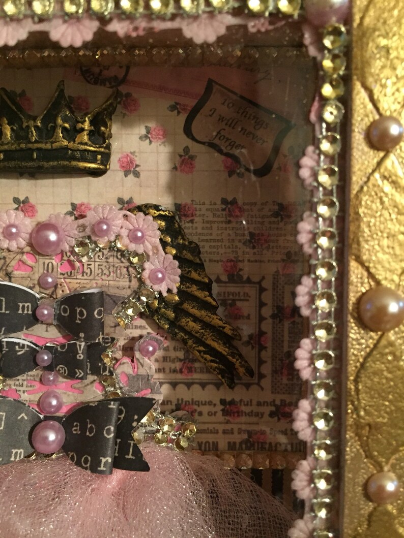 May include: A pink and gold shadow box with a crown, wings, and ribbons. The box is decorated with pink flowers and pearls. The text "10 things I will never forget" is visible in the box.