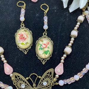 May include: A vintage jewellery set featuring a butterfly pendant necklace, earrings, and a bracelet. The necklace has a filigree butterfly pendant with clear rhinestones. The earrings and pendant feature oval-shaped images of flowers. The set is in shades of pink and gold.