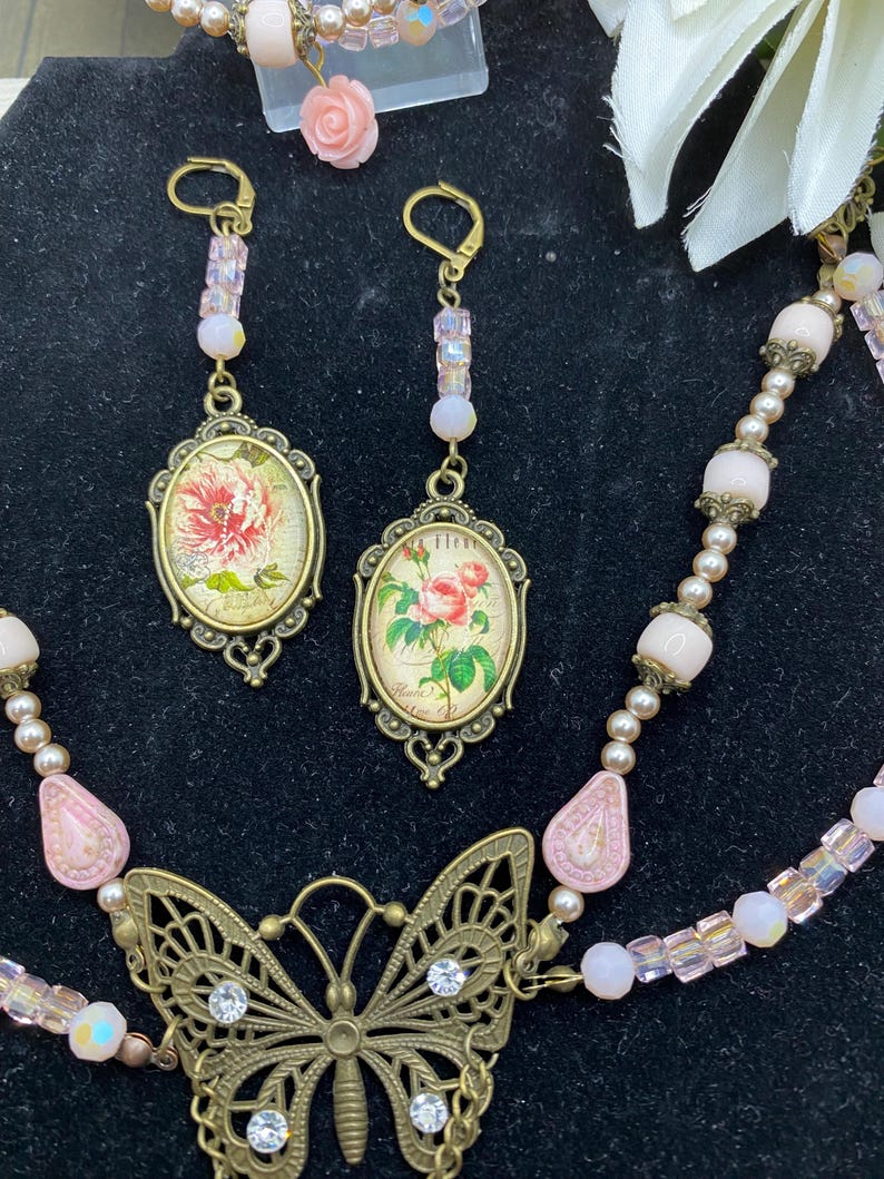 May include: A vintage jewellery set featuring a butterfly pendant necklace, earrings, and a bracelet. The necklace has a filigree butterfly pendant with clear rhinestones. The earrings and pendant feature oval-shaped images of flowers. The set is in shades of pink and gold.