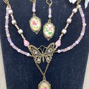 May include: A vintage jewellery set comprising a necklace and earrings. The necklace features a butterfly pendant and a floral oval pendant. The earrings have matching floral oval pendants. The set is decorated with pink beads and crystals, with gold-toned metal accents.
