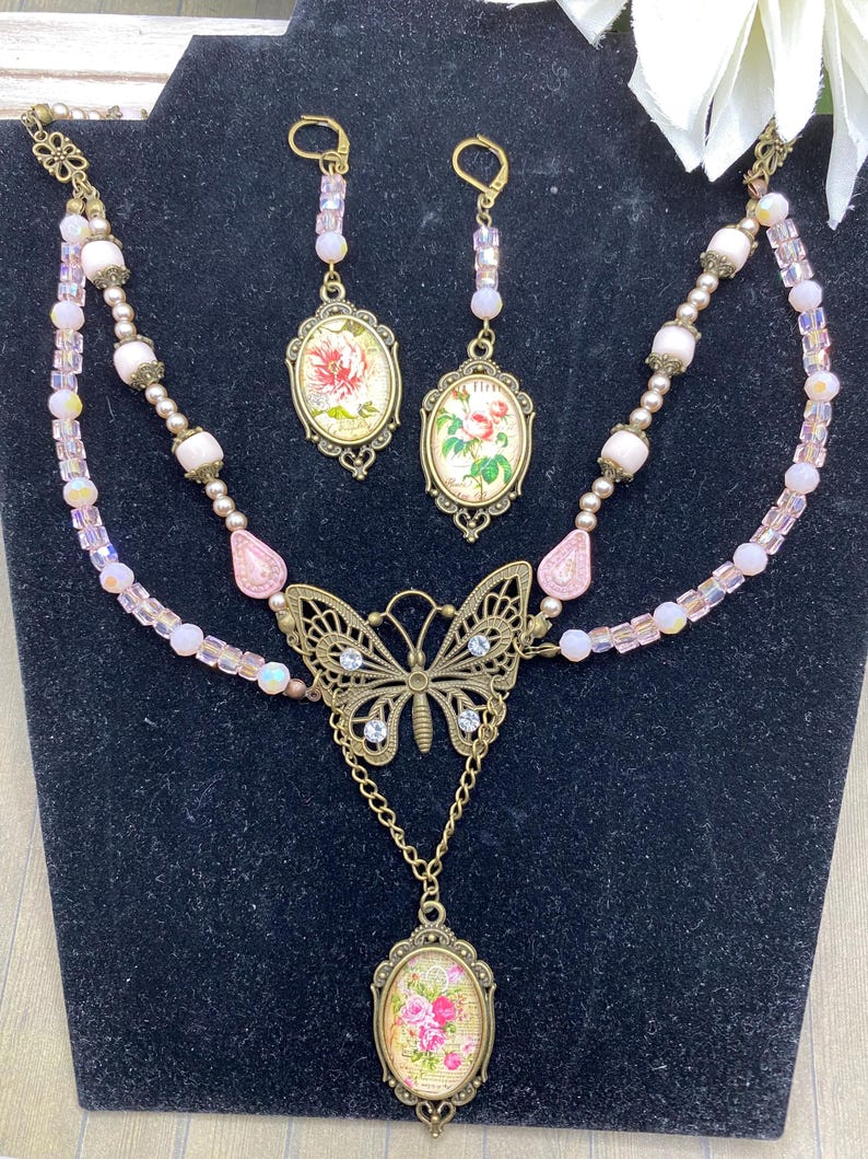 May include: A vintage jewellery set comprising a necklace and earrings. The necklace features a butterfly pendant and a floral oval pendant. The earrings have matching floral oval pendants. The set is decorated with pink beads and crystals, with gold-toned metal accents.