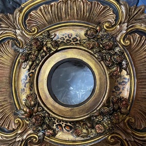 May include: Ornate, gold-colored mirror frame with a circular center. The frame features intricate floral and leaf designs, with a leopard print pattern around the center. The mirror is surrounded by sculpted roses.