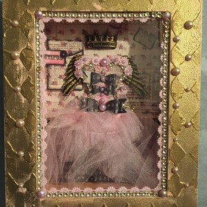 May include: A framed shadow box with a pink tulle tutu, a crown, and wings. The box is decorated with gold paint, rhinestones, and pearls. The words "Untiful" and "I'm a princess" are visible inside the box.