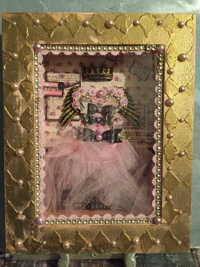 May include: A framed shadow box with a pink tulle tutu, a crown, and wings. The box is decorated with gold paint, rhinestones, and pearls. The words "Untiful" and "I'm a princess" are visible inside the box.