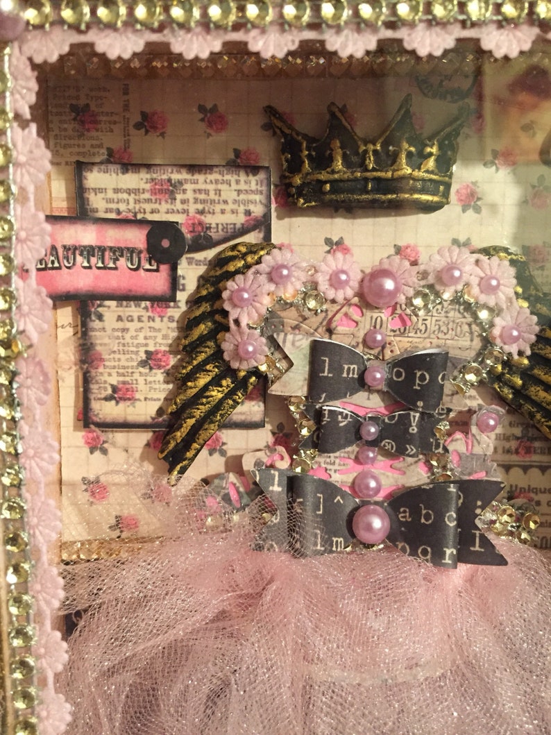 May include: A pink tulle tutu with black and white ribbon bows and a gold crown. The tutu is decorated with pink and white flowers and pearls. The text "1 mopo" and "abc" is visible on the ribbons.
