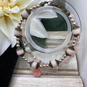 May include: A double-strand bracelet with pale pink beads, small pearls, and decorative metal elements. The bracelet includes a small pink rose charm. The bracelet is displayed on a clear glass surface.