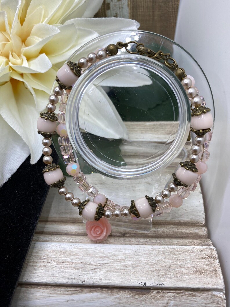 May include: A double-strand bracelet with pale pink beads, small pearls, and decorative metal elements. The bracelet includes a small pink rose charm. The bracelet is displayed on a clear glass surface.
