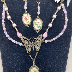 May include: A vintage jewellery set comprising a necklace and matching earrings. The necklace features a butterfly pendant and a floral oval pendant. The earrings have oval pendants with floral designs. The set is decorated with pink beads and crystals.