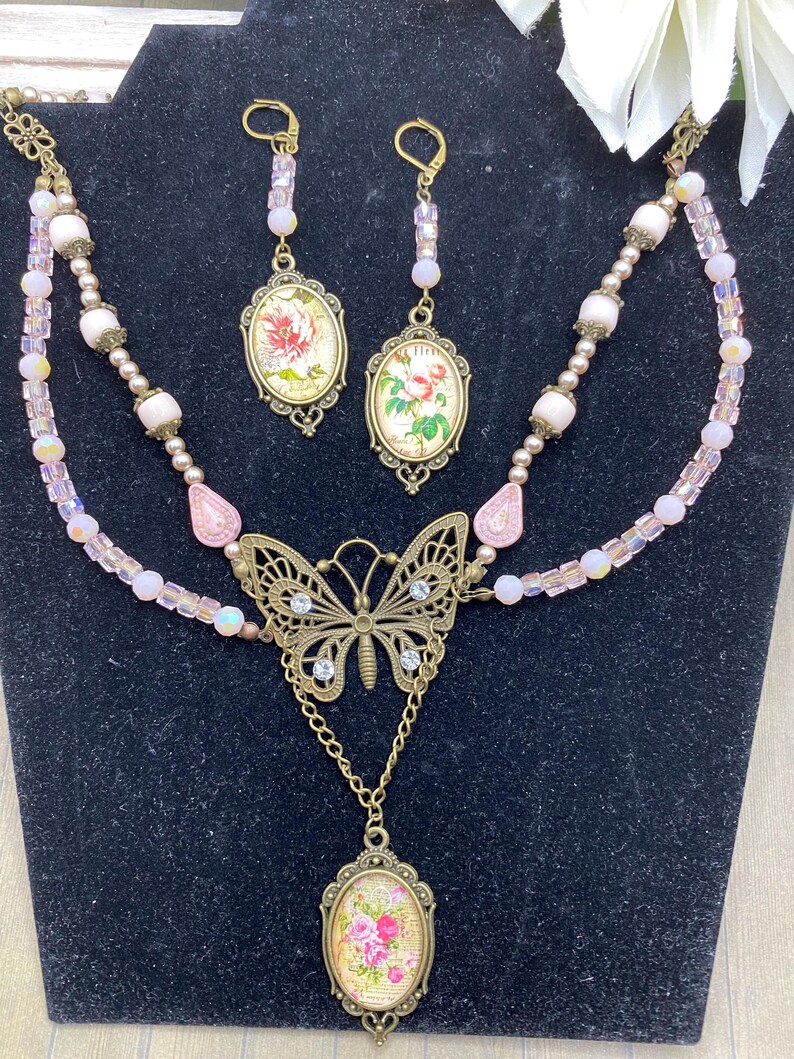 May include: A vintage jewellery set comprising a necklace and matching earrings. The necklace features a butterfly pendant and a floral oval pendant. The earrings have oval pendants with floral designs. The set is decorated with pink beads and crystals.