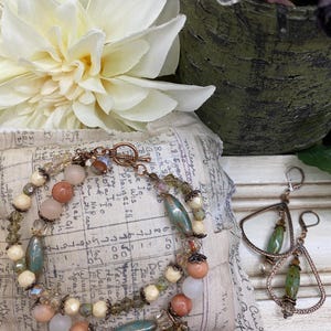May include: A beaded bracelet and matching earrings. The bracelet features peach, cream, and teal beads with a copper clasp. The earrings have a copper frame with a teal bead center. The jewelry is displayed on a vintage fabric.