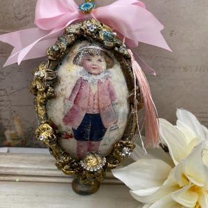 May include: An oval-shaped, vintage-style ornament featuring a child in a pink coat and blue shorts. The ornament has a gold-colored frame with floral detailing, embellished with rhinestones and a light pink bow. A small tassel hangs from the bottom.