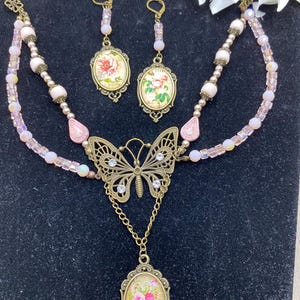 May include: A vintage jewellery set comprising a necklace, earrings, and bracelet. The necklace features a butterfly pendant and a floral pendant. The earrings and pendants have floral designs. The set is in shades of pink and gold.