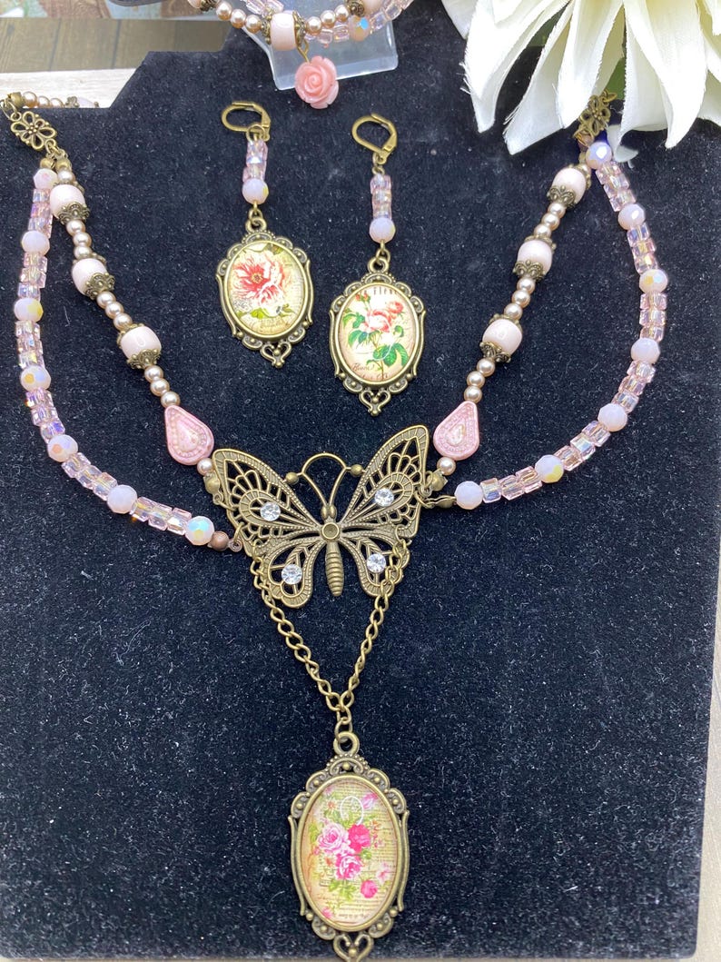 May include: A vintage jewellery set comprising a necklace, earrings, and bracelet. The necklace features a butterfly pendant and a floral pendant. The earrings and pendants have floral designs. The set is in shades of pink and gold.