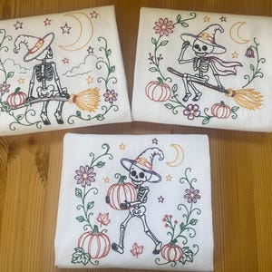 May include: Three white fabric squares featuring colorful machine embroidery designs of skeletons. Each square displays a skeleton wearing a witch hat, surrounded by floral vines, pumpkins, stars, and a crescent moon. The embroidery uses green, orange, purple, and yellow thread.