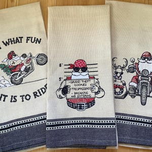 May include: Three kitchen towels with embroidered designs featuring Santa Claus riding a motorcycle. The first towel says "Oh What Fun It Is To Ride". The second towel features Santa Claus with a mugshot and the text "Grand Theft Gookies, Trespassing, Breaking and Entering". The third towel features Santa Claus riding a motorcycle with a sidecar.