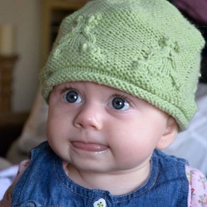 May include: A baby wearing a green knitted hat with a textured pattern and a blue denim pinafore dress.