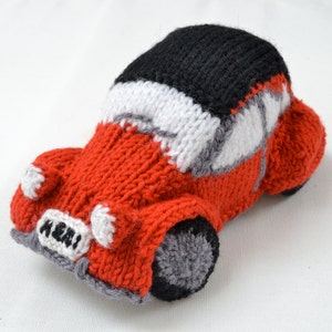 Peut inclure: Une voiture jouet en tricot rouge avec un toit en tricot noir, des détails en tricot blanc et une roue en tricot gris. La voiture a une plaque d'immatriculation en tricot blanc qui dit "A121".