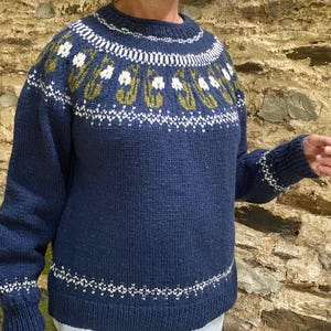 Pode incluir: Um suéter de malha azul marinho com decote redondo. O suéter apresenta um padrão floral branco e verde-oliva no peito e na parte superior das mangas, com detalhes brancos na gola, punhos e bainha. O suéter é feito de um fio espesso e texturizado.