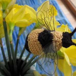 May include: A crocheted bee with black and brown stripes and clear wings sits among yellow daffodils.