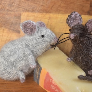 May include: Two knitted mice, one grey and one brown, are sitting on a block of cheese. The grey mouse has a long tail and is looking at the camera. The brown mouse has short ears and is looking to the right. The cheese is wrapped in red and white paper with the words "First Press from" printed on it.