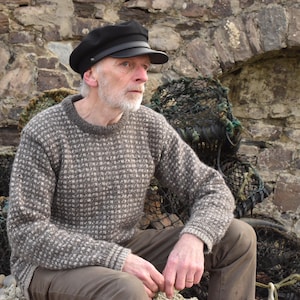May include: A man wearing a brown and white patterned jumper and a black cap sits in front of a stone wall. He is wearing brown trousers and has a white beard.