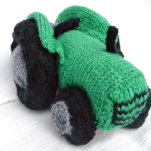 May include: A handmade knitted toy tractor in shades of green, black, and gray. The toy features a detailed design with wheels, a cab, and other tractor elements. The soft, textured appearance suggests a cozy, child-friendly item.