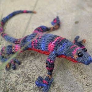 May include: A knitted lizard toy with a grey, blue, pink and orange colour scheme. The lizard has black eyes and a long tail.