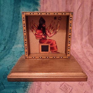 May include: A rectangular wooden display frame with a tan and black geometric border, featuring an illustration of a seated figure with wings, a beard, and a red robe. The frame is mounted on a solid, smooth, light brown wooden base stand.