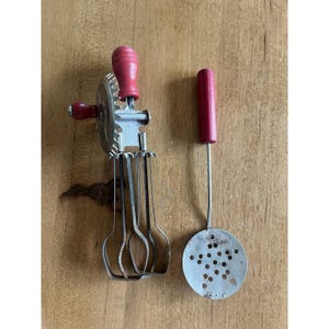 May include: Vintage kitchen utensils set. Includes a hand-crank egg beater with a red wooden handle and a slotted spoon with a red wooden handle. The metal utensils show signs of wear.