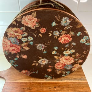 May include: A circular brown hat box adorned with a floral design in shades of pink, peach, and blue. It features a rope handle and copper-toned trim. The box is placed on a wooden chopping board.