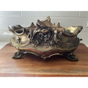 May include: An ornate, metallic, brass-toned centerpiece bowl featuring intricate relief carvings of floral motifs and scrolling edges. The vessel is supported by four curved feet and displays a weathered, darkened patina across its textured surface.