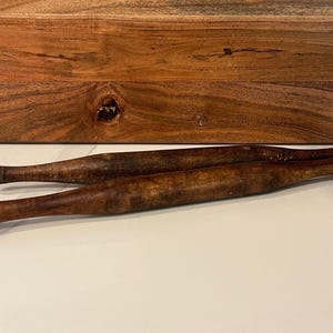 May include: Two wooden rolling pins, one on top of the other, on a white surface. The rolling pins are brown and have a natural wood finish.