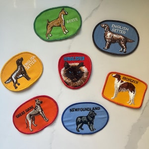 May include: Embroidered patches featuring various dog breeds and a Himalayan cat. Each patch has a different colored oval background with the breed's name and an image of the animal. Breeds include Boxer, English Setter, Great Dane, and Newfoundland.