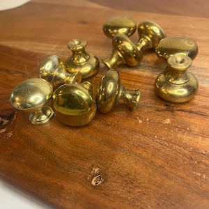 May include: A collection of brass cabinet knobs with a polished finish. The knobs have a rounded shape with a small base and a threaded post for attachment. They are arranged on a wooden surface.
