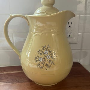 May include: A pale yellow vintage pitcher with a matching lid and handle. The pitcher features a floral design with blue and white flowers and gray leaves. The pitcher is sitting on a wooden surface.