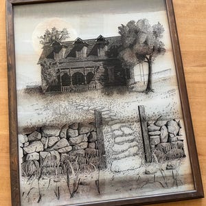 以下が含まれることがあります： 額縁に入った、黒と白のペンとインクの素朴な家の絵。前景には石垣があります。家にはポーチとドーマー窓があり、家の右側にはブランコのある木があります。