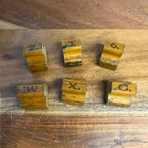 May include: Six small, weathered wooden blocks with numbers and letters printed on them. The blocks are various shades of brown and yellow, with the numbers and letters in black. The blocks are arranged on a wooden surface.