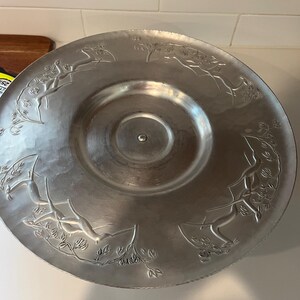 Puede incluir: Una fuente redonda plateada con una textura martillada. El borde presenta un diseño en relieve de ciervos saltando entre follaje. La fuente tiene un centro ligeramente cóncavo y un pequeño pomo central.