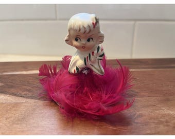 May include: A ceramic figurine featuring a stylized head with blonde hair and painted facial features, attached to a base made of vibrant magenta feathers. The figure has a white collar with green and red accents and a small clear rhinestone detail.