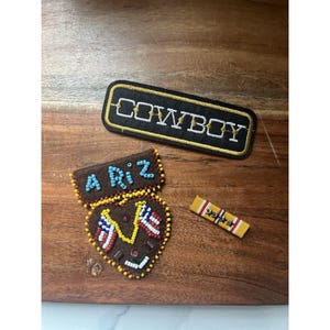 May include: A rectangular black patch with gold trim and the white text COWBOY, a brown heart-shaped beaded patch with the light blue text A RiZ and two American flag motifs, and a small rectangular yellow ribbon bar with red stripes and stars.