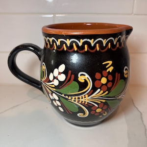 Puede incluir: Jarra de cerámica negra con asa y boquilla, decorada con coloridos diseños florales y de vid. El borde está adornado con una banda marrón y un patrón ondulado blanco, amarillo y rojo. Una pieza encantadora para servir bebidas.