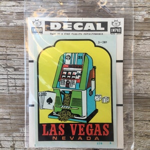 May include: A decal featuring a vintage slot machine with the text "LAS VEGAS NEVADA". The decal is in a clear plastic sleeve. The slot machine is green, blue, and red, with a yellow background. Includes playing cards and dice.