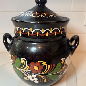 Puede incluir: Olla de cerámica negra con tapa y dos asas, adornada con diseños florales. La olla tiene una tapa con un pomo y está decorada con patrones florales blancos, amarillos, rojos y verdes. La olla probablemente se usa para cocinar o servir.