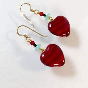 Puede incluir: Un par de pendientes de corazón dorados con cuentas de vidrio rojas y verdes. Las cuentas con forma de corazón tienen un patrón de espiral rojo y blanco.